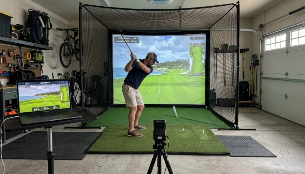 A home golf simulator setup in a garage with a hitting mat, impact screen, and laptop displaying a virtual golf course