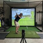 A home golf simulator setup in a garage with a hitting mat, impact screen, and laptop displaying a virtual golf course