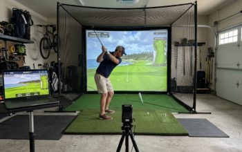 A home golf simulator setup in a garage with a hitting mat, impact screen, and laptop displaying a virtual golf course